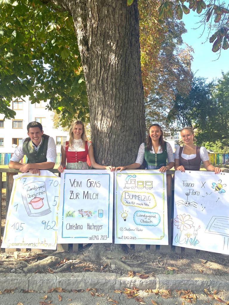 Vier junge Menschen in Tracht stehen unter einem Baum. Jede/r hält ein Poster mit ihrer Projektidee in der Hand